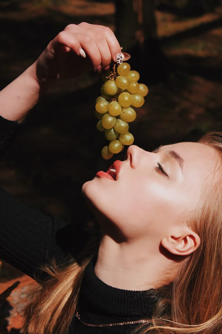 a woman holding grapes up to her face with one hand and the other eye closed