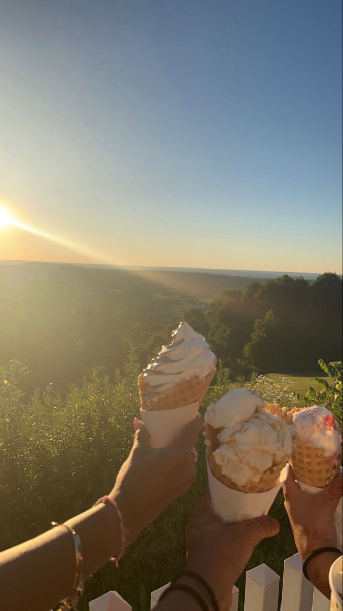 two people holding ice cream cones in their hands with the sun setting behind them,