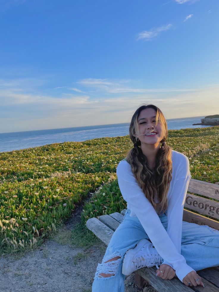 a young woman sitting on top of a wooden bench next to the ocean and grass