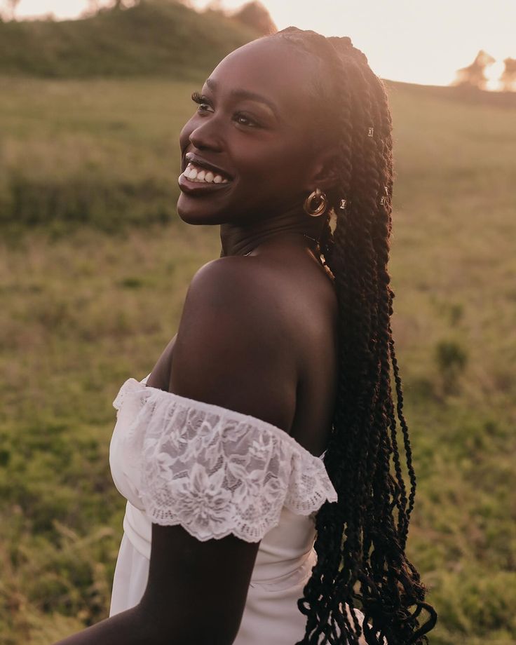 a woman standing in a field smiling at the camera with her hair pulled back and wearing a white top