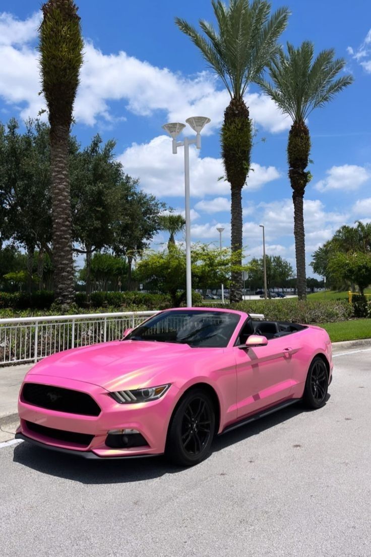 a pink mustang parked in a parking lot next to palm trees and street lights with blue skies behind it