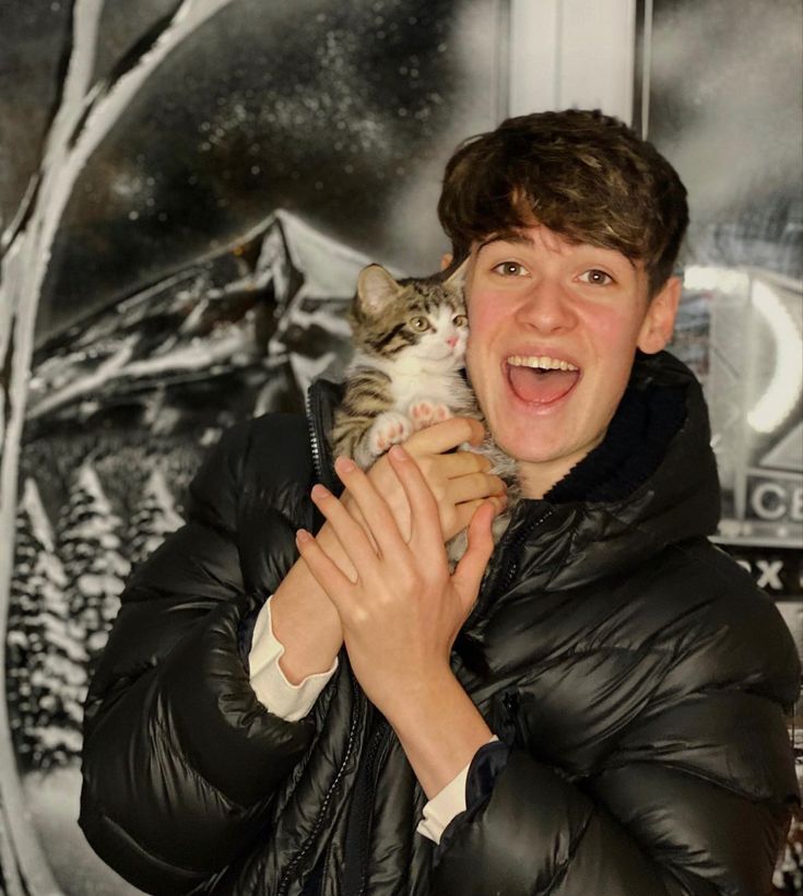 a young man holding a cat in his arms and smiling at the camera while wearing a black jacket