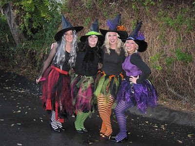 four women dressed up in halloween costumes posing for the camera on a road with trees and bushes behind them