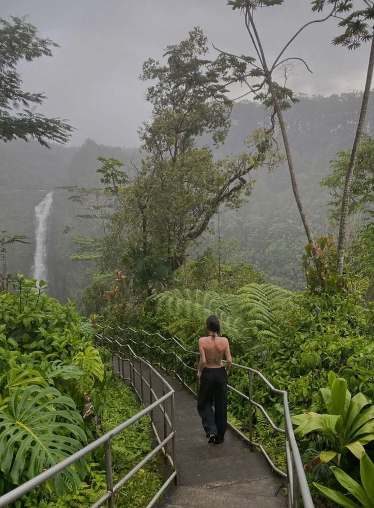 a woman walking up stairs in front of a waterfall with lush vegetation and trees on both sides