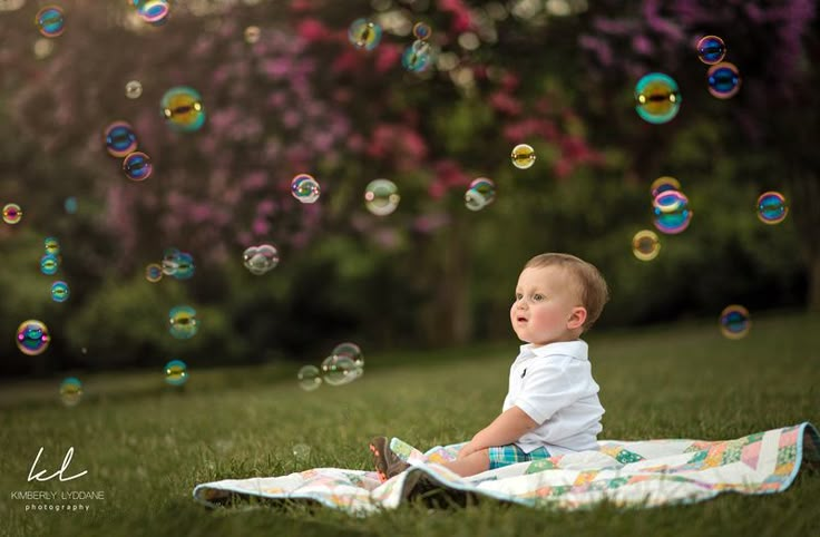 a toddler sitting on a blanket in the grass with soap bubbles floating around him