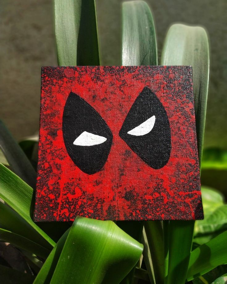 a red and black square with white eyes on top of some green plants in front of a brick wall