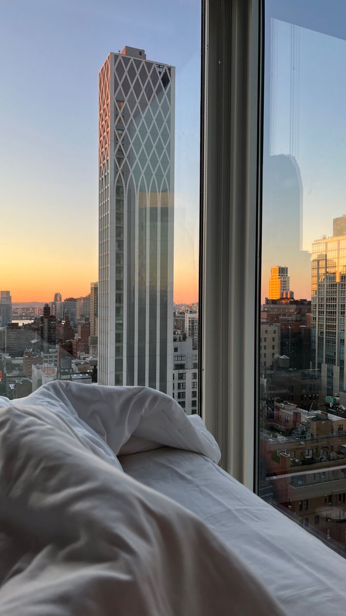 an unmade bed in front of a large window overlooking the city skyline at sunset