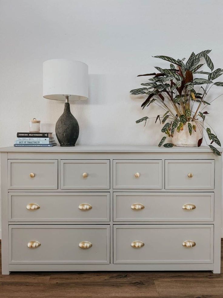 a white dresser with gold handles and drawers in a room next to a lamp on top of a wooden floor