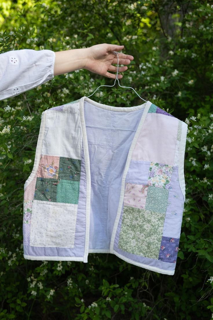 a person holding up a white shirt with patchwork on it in front of some bushes