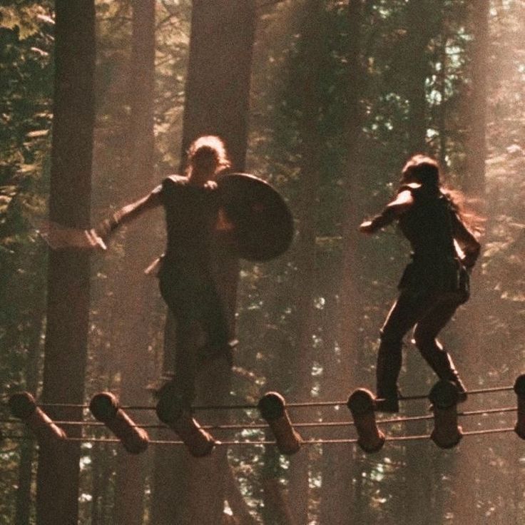 two people are walking across a rope in the woods, one is carrying a shield