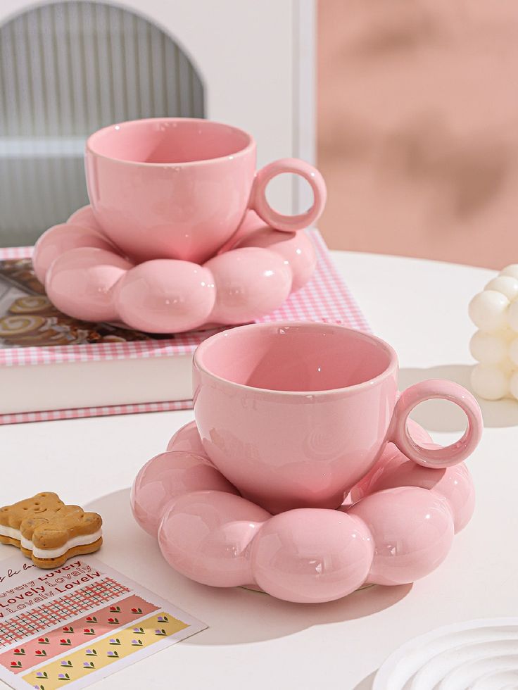 two pink cups and saucers sitting on a table with cookies in front of them