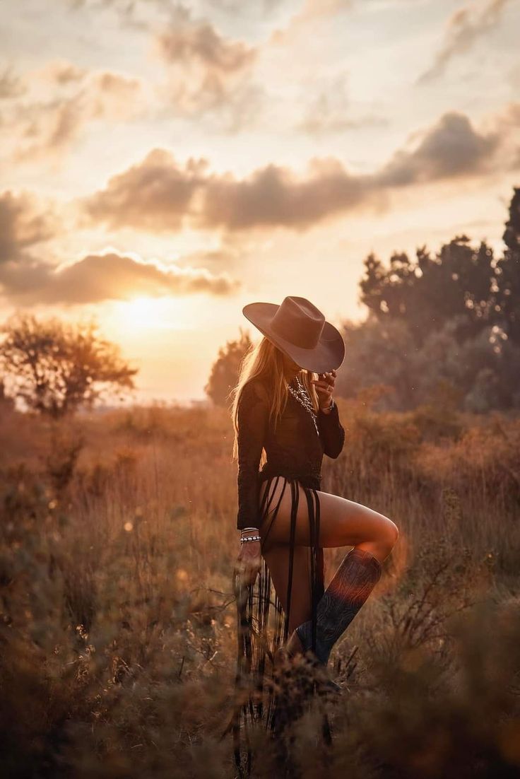 a woman wearing a cowboy hat standing in tall grass with the sun setting behind her