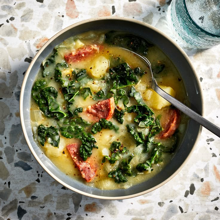 a bowl of soup with spinach, ham and cheese on the table next to a glass of water