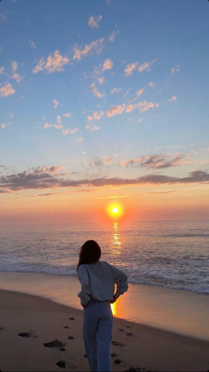 a person standing on top of a beach next to the ocean at sunset with footprints in the sand