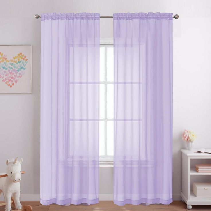 a white teddy bear sitting in front of a window with purple sheer curtains on it