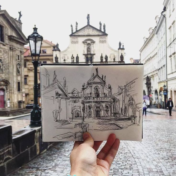 a hand holding up a drawing in front of a building on a brick street with people walking by