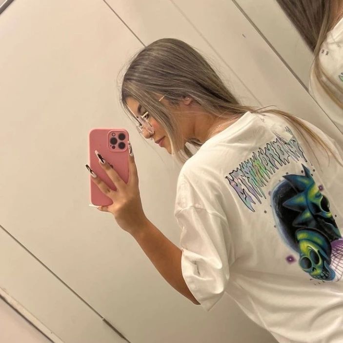 a girl looking at her cell phone while standing in front of a mirror wearing a t - shirt