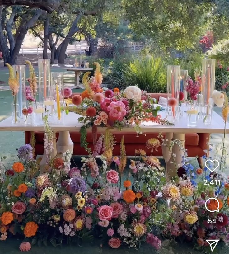 a table with flowers and candles on it in the middle of a park area, surrounded by trees