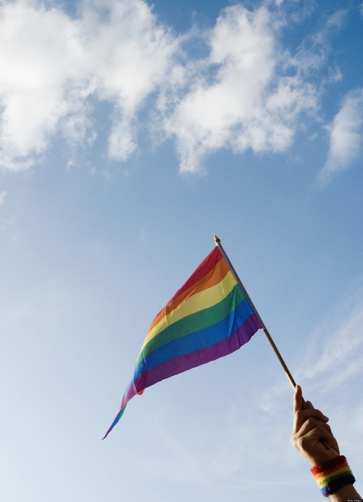 a hand holding a rainbow flag in the air with clouds behind it and blue sky