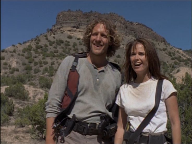 a man and woman standing next to each other in front of a mountain with trees