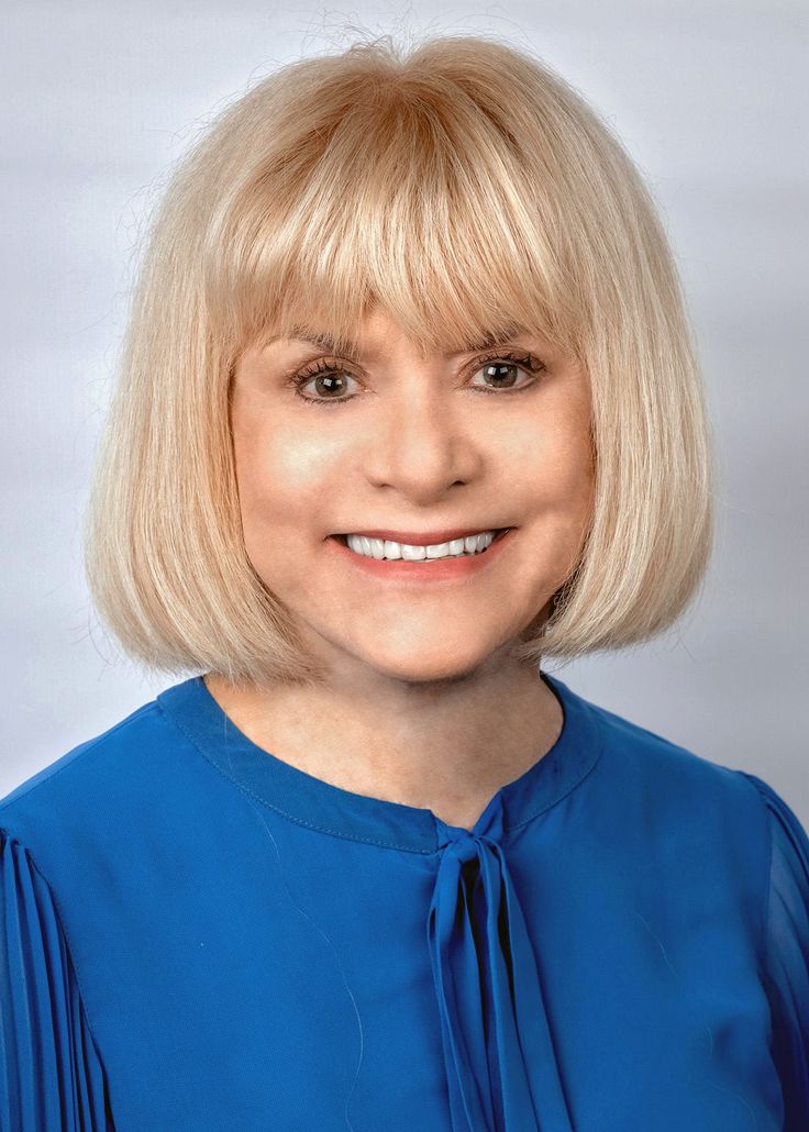 a woman with blonde hair wearing a blue blouse and smiling at the camera while standing in front of a white background