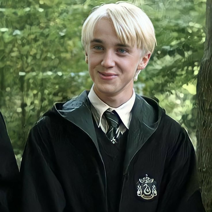 a young man with blonde hair wearing a black jacket and tie in front of trees