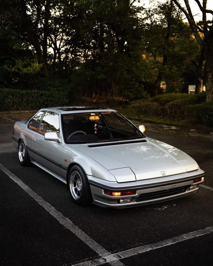 a silver car parked in a parking lot next to some trees and bushes at sunset