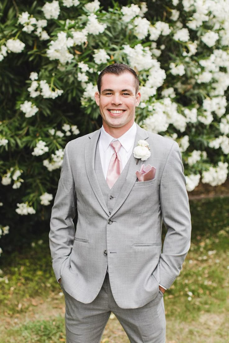 a man in a gray suit and pink tie standing next to a bush with white flowers