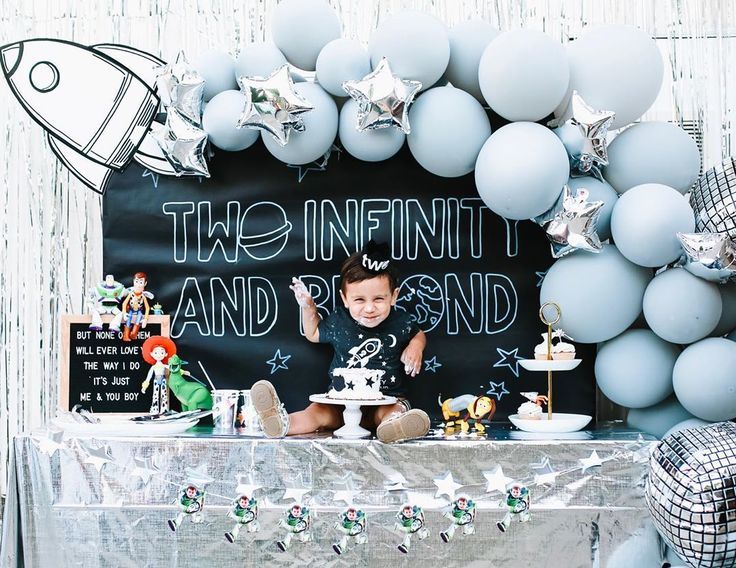 a little boy sitting in front of a table with cake and balloons on it's side