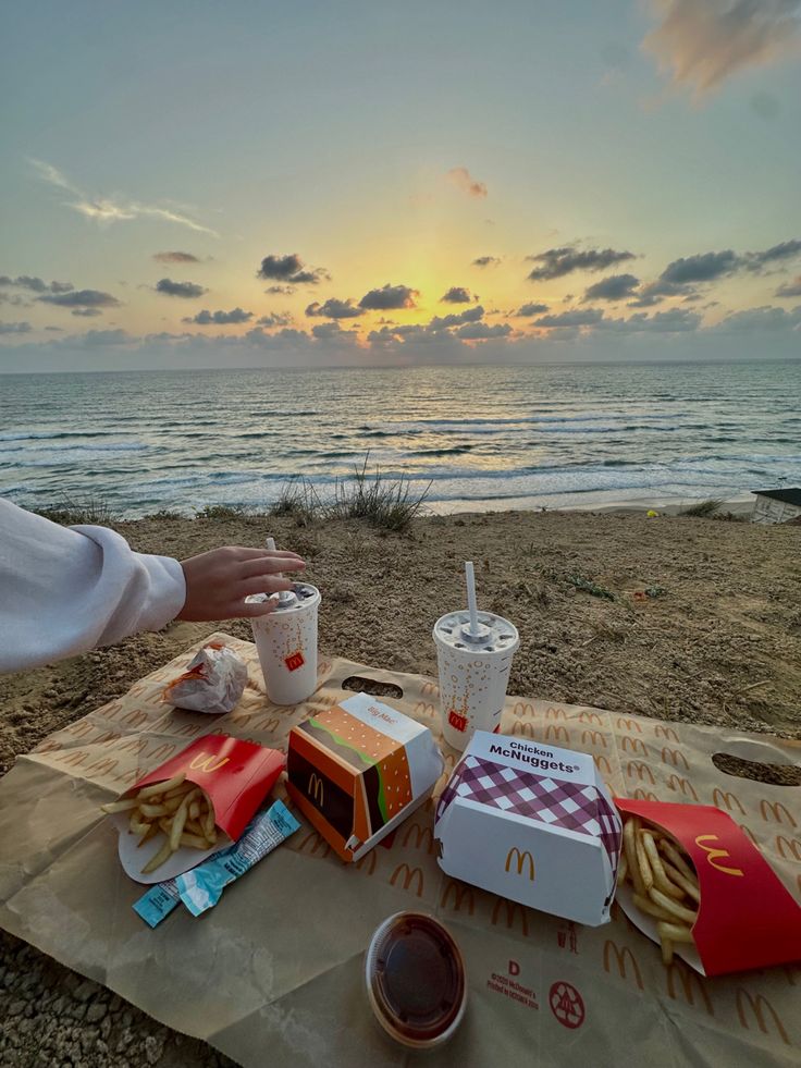 a person sitting at a picnic table on the beach with food and drinks in front of them