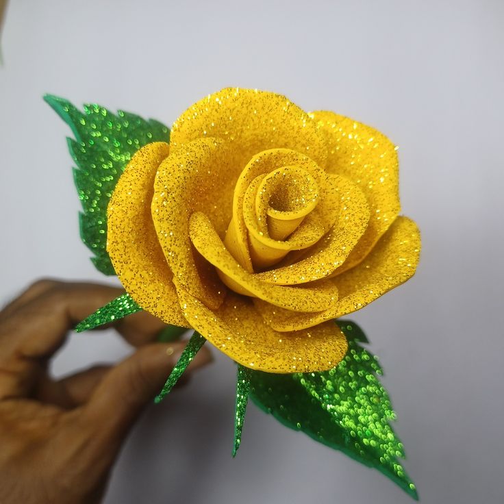 a yellow rose with green leaves is being held up by someone's hand on a white surface