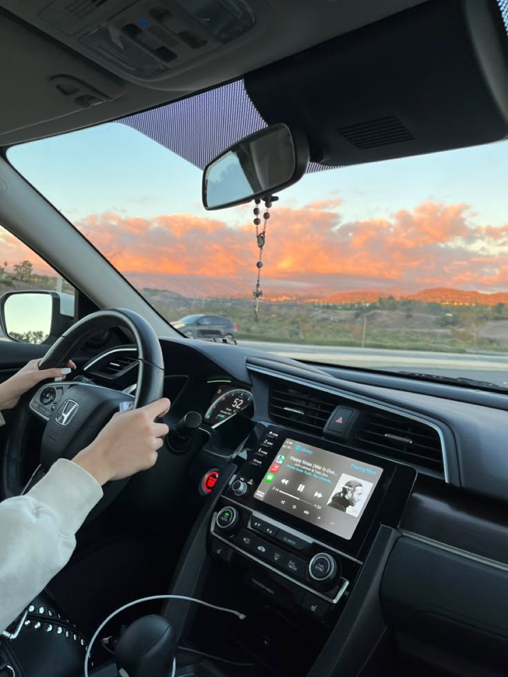 a woman is driving her car with the sun setting in the back ground and pink clouds