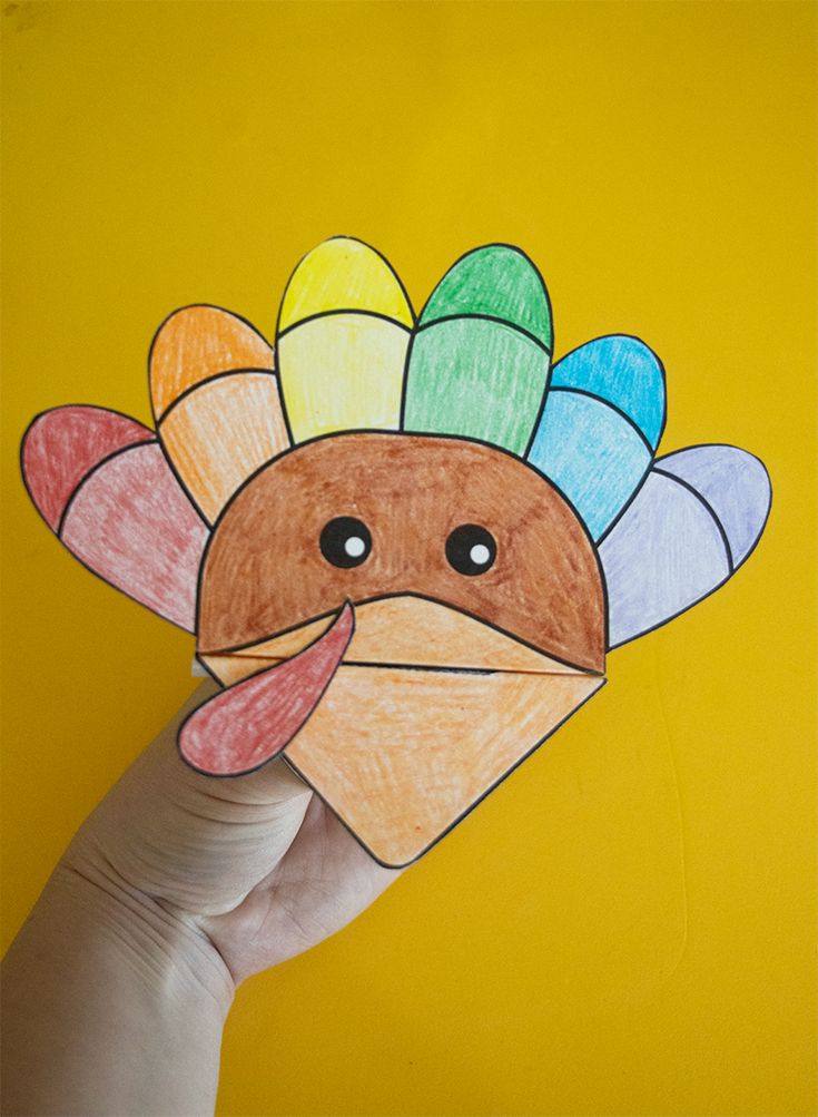 a hand holding up a paper turkey with colored feathers on it's head and eyes