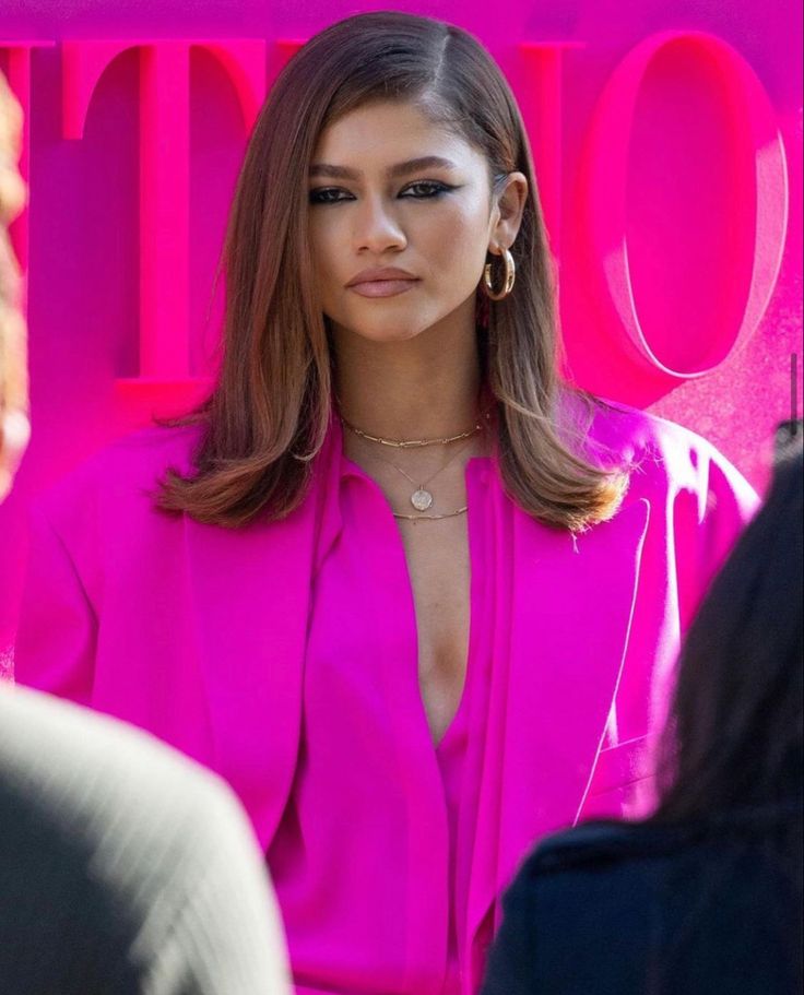 a woman with long hair wearing a pink shirt and gold hoop earrings standing in front of a neon sign