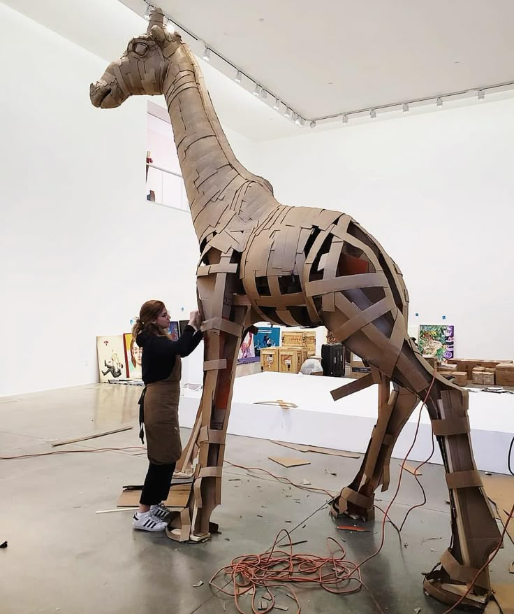 a woman standing next to a wooden sculpture of a giraffe with strips of tape on it's body