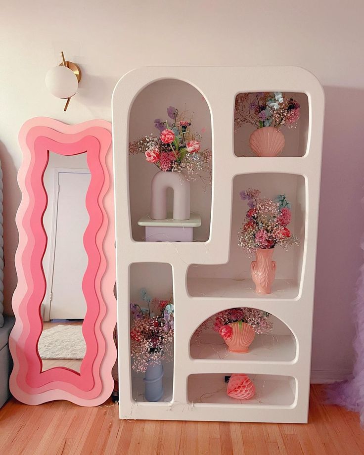 a white shelf with vases and flowers on it in front of a pink mirror