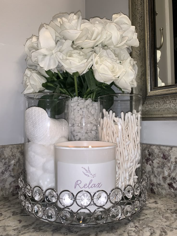 white flowers and candles sit in a glass container on a marble countertop next to a mirror