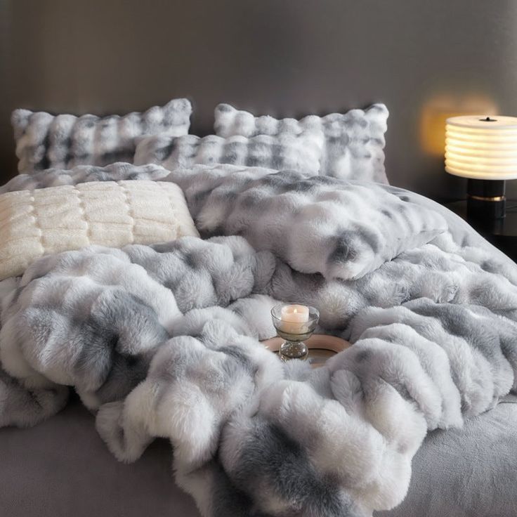 a bed covered in grey and white furs next to a night stand with a lamp on it