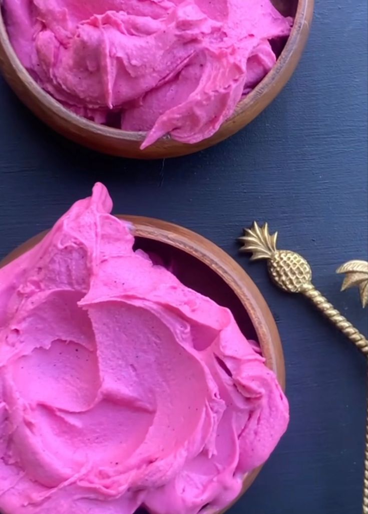 two wooden bowls filled with pink frosting next to a gold pineapple decoration on a blue surface