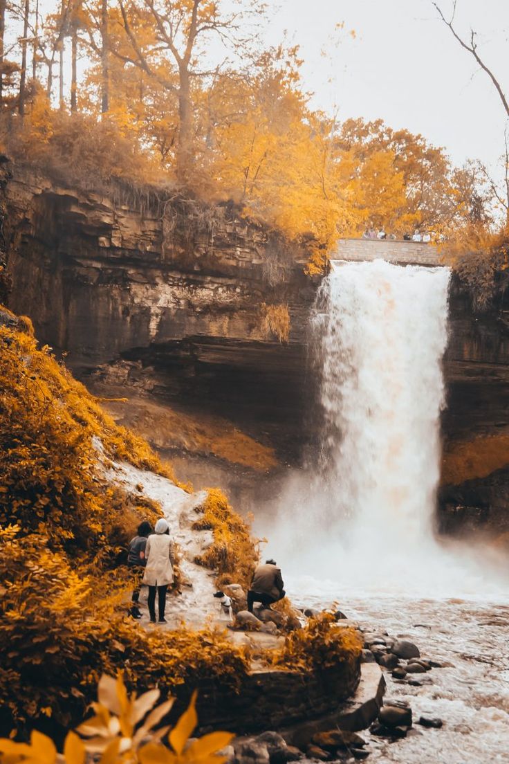 a man standing in front of a waterfall with fall foliage around him and his back to the camera