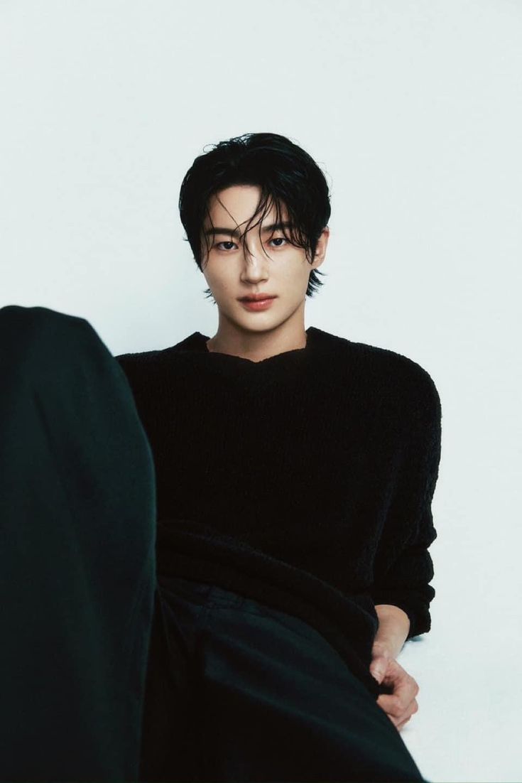 a young man sitting on top of a black chair next to a white wall with his hands in his pockets