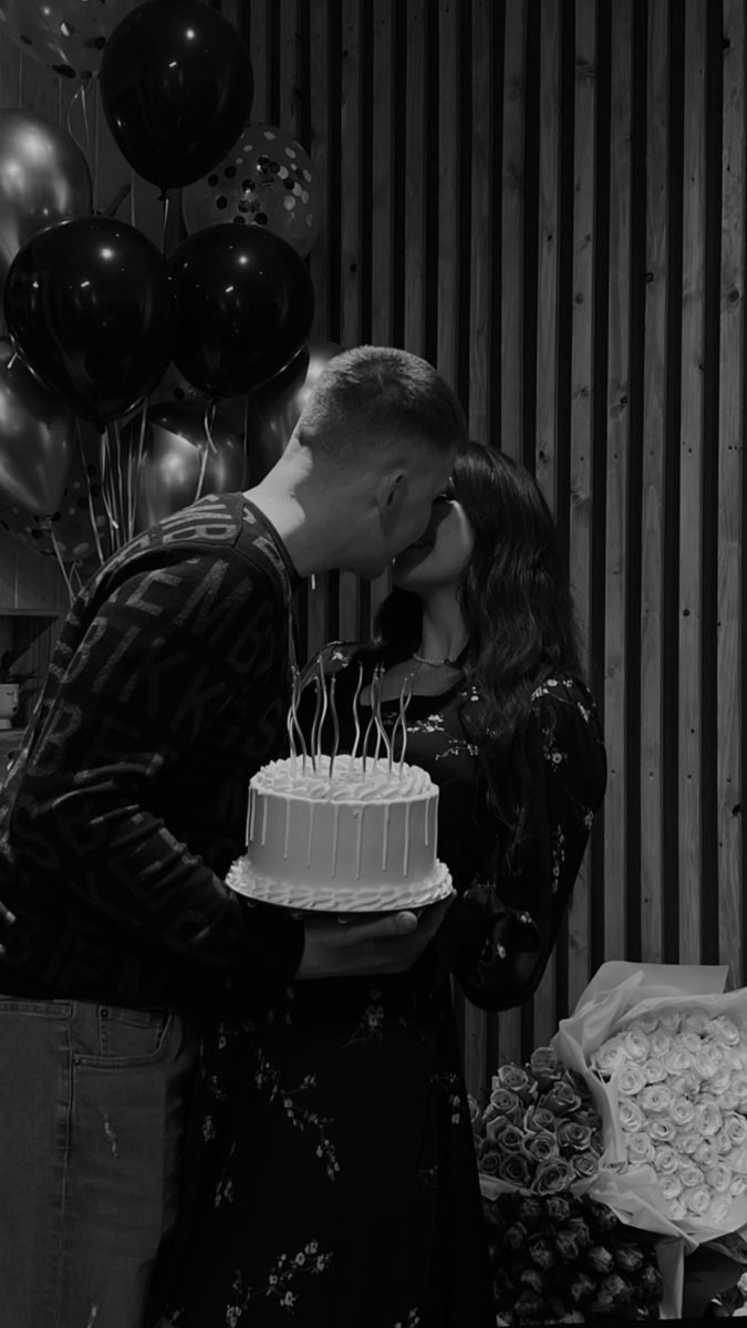 a man and woman kissing in front of a cake with candles on it, surrounded by balloons