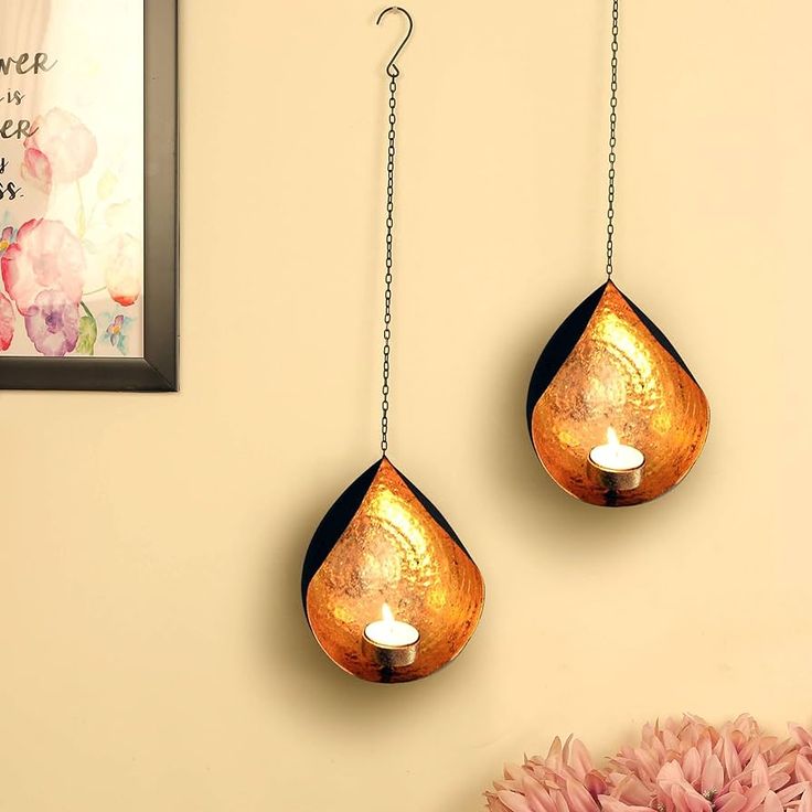 two hanging tea light holders on a wall with pink flowers in the corner next to it