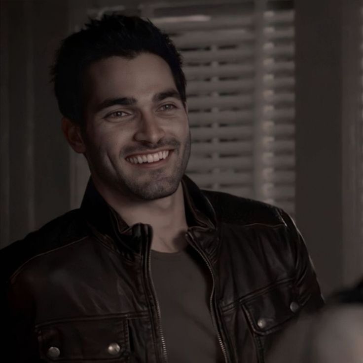 a man in a leather jacket smiling at the camera while standing next to a window