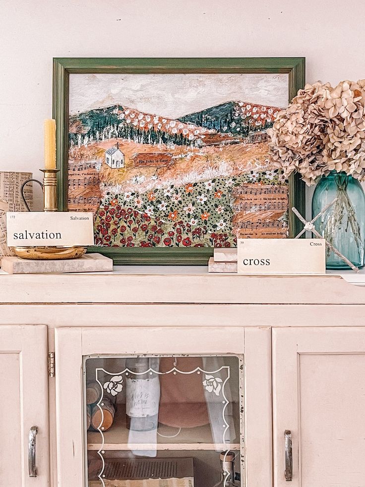 a painting is on the wall above a china cabinet with flowers and other items in it