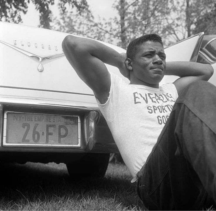 a black and white photo of a man sitting in front of a car with his hand on his head