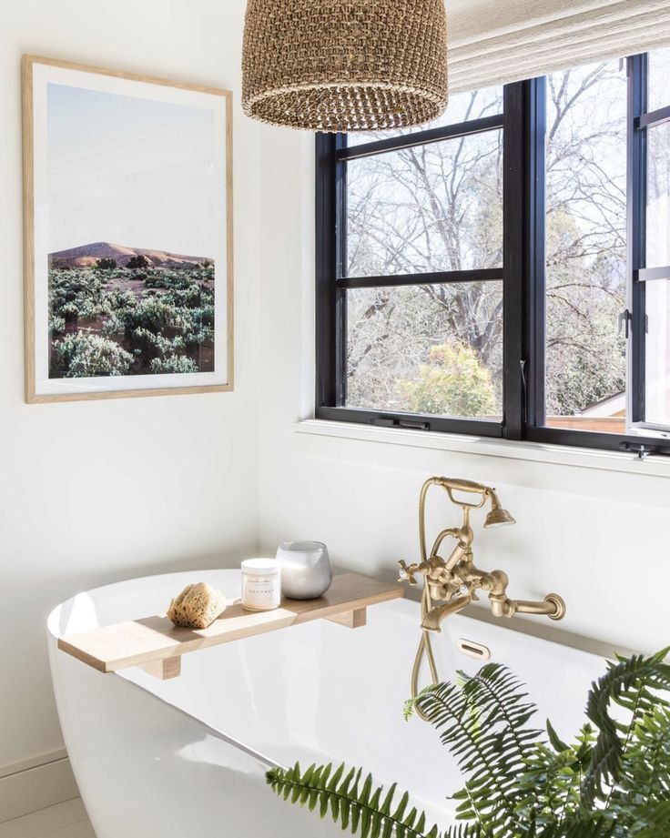 a white bath tub sitting under a window next to a potted plant and framed photograph