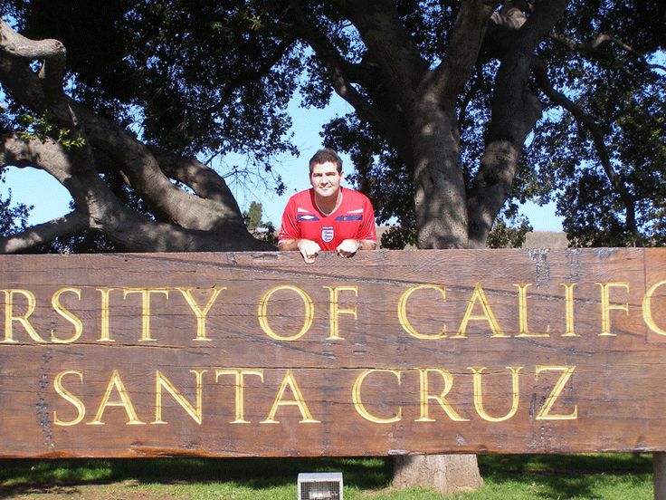 a man sitting on top of a wooden sign in front of a tree with the words university of california santa cruz