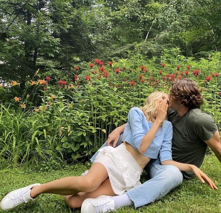 a man and woman sitting on the ground in front of some flowers, one is kissing the other's cheek