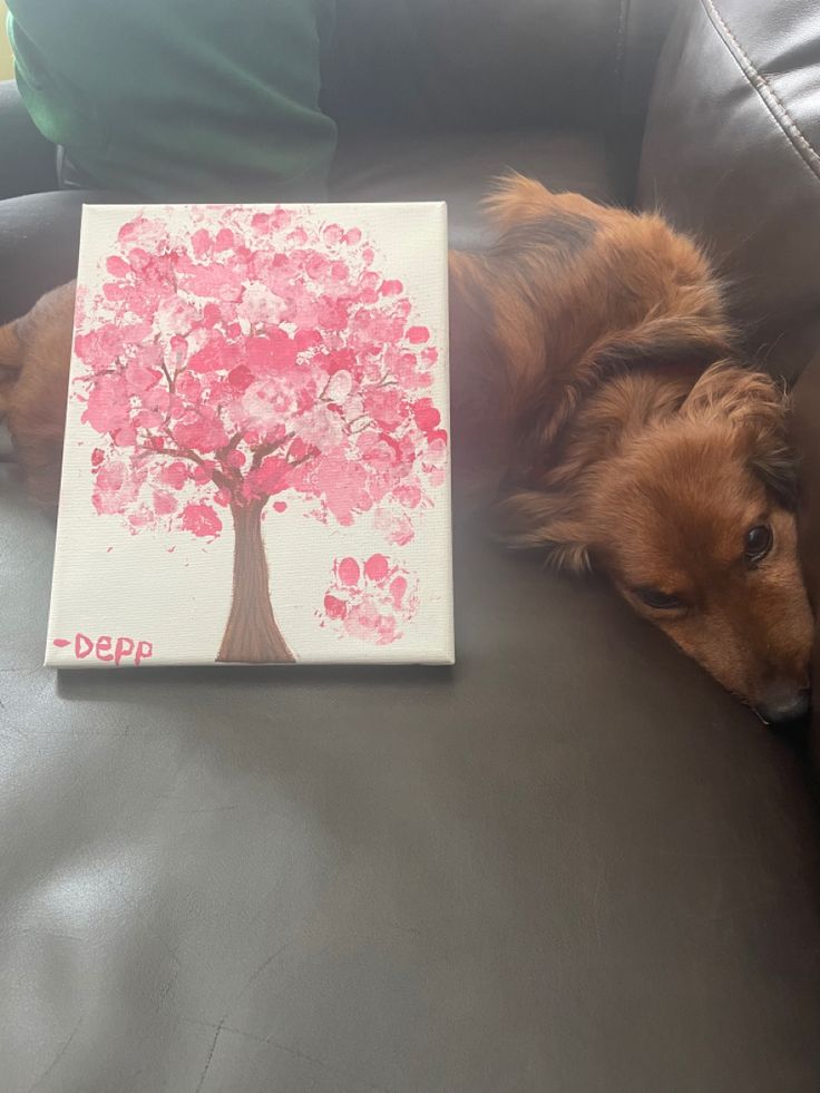 a brown dog laying on top of a couch next to a book with a tree painted on it